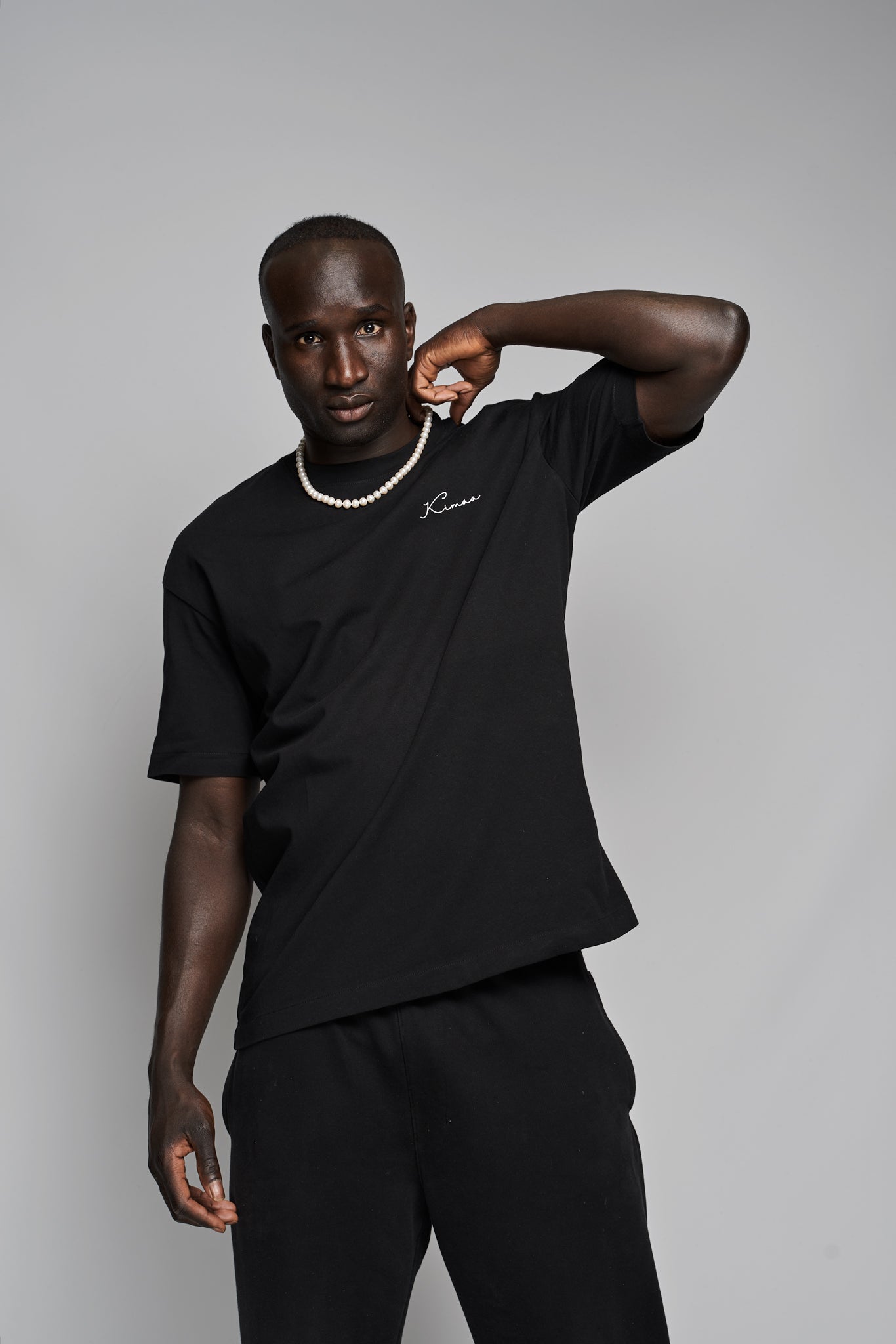 Model wearing Suutoo black organic cotton T-shirt by Kimaa, holding hand up with necklace, expressive Afro-Nordic style.